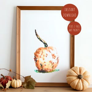 May include: Watercolor print of a pumpkin in a wooden frame. The pumpkin is orange with yellow highlights and a green stem. The print is labeled "INSTANT DOWNLOAD" and "PLUS 2 FREE ART PRINTS". Two pumpkins and a small gourd are in the foreground.