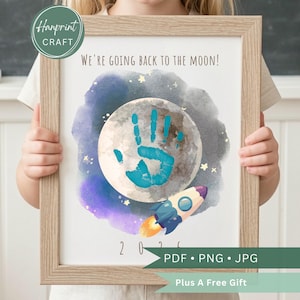 May include: A framed print with a watercolor design featuring a moon with a blue handprint, a rocket, and the text "WE'RE GOING BACK TO THE MOON!". The print also includes the text "202" and "PDF • PNG • JPG".