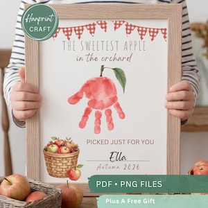 May include: A framed print featuring a red handprint shaped like an apple, red gingham bunting, and a basket of apples. The text reads: THE SWEETEST APPLE in the orchard, PICKED JUST FOR YOU, Ella, Autumn 2026. The frame is light wood.