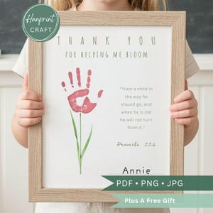 Puede incluir: Una impresión enmarcada con un diseño de flor de huella de mano. El texto dice "THANK YOU FOR HELPING ME BLOOM" e incluye un versículo bíblico. La impresión está en un marco de madera marrón claro e incluye el nombre "Annie".