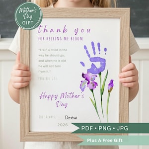 Puede incluir: Un marco con un marco de madera clara. La impresión tiene una huella de mano morada y flores de iris de acuarela. El texto incluye "Gracias", "Feliz Día de la Madre" y "Drew 2026". Un regalo del Día de la Madre.