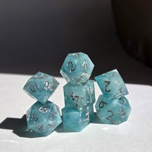 Könnte beinhalten: Ein Satz von sieben durchscheinenden blauen Würfeln mit silbernen Zahlen. Die Würfel haben verschiedene polyedrische Formen, darunter ein d20, d12, d10, d8 und d6. Die Würfel haben ein wolkiges, marmoriertes Aussehen und sind auf einer weißen Oberfläche angeordnet.