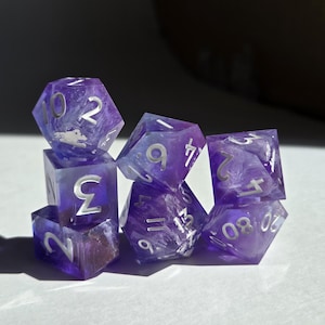 Pode incluir: Um conjunto de sete dados de resina roxa com numeração prateada. Os dados têm um efeito marmorizado, com uma mistura de roxo translúcido e opaco. Os dados estão empilhados, alguns parcialmente escondidos. Para jogos de tabuleiro.