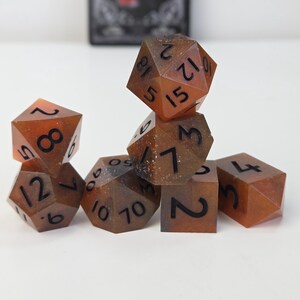 Op de afbeelding: Een set van zeven polyhedrale dobbelstenen in bruin- en amberkleuren met zwarte cijfers. De dobbelstenen zijn in een kleine stapel gerangschikt. Deze dobbelstenen zijn voor tabletop games. De afmetingen zijn in centimeters.