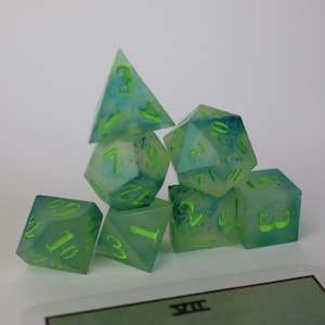 Pode incluir: Um conjunto de sete dados translúcidos em várias formas geométricas. Os dados são uma mistura de verde claro e azul com números verdes brilhantes. Os dados estão empilhados, com um cartão em primeiro plano.