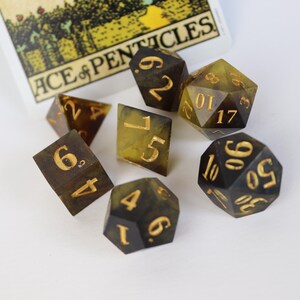Pode incluir: Um conjunto de sete dados poliédricos em tons de castanho e amarelo com numeração dourada, dispostos ao redor de uma carta de tarô. Os dados são para jogos de tabuleiro. A carta diz "ACE of PENTACLES".