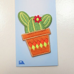 May include: An embroidered iron-on patch featuring a green cactus with a small red flower in an orange pot. The pot has a diamond pattern in orange and light green. The patch is attached to a light blue card with a small iron icon at the bottom.