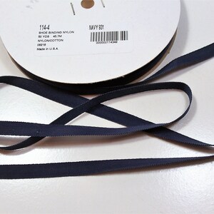 May include: A spool of navy blue shoe binding nylon ribbon. The label reads "114-4 SHOE BINDING NYLON 50 YDS 45.7M NYLON/COTTON 08218 Made in U.S.A. NAVY 931". The ribbon is laid out in a looping pattern.