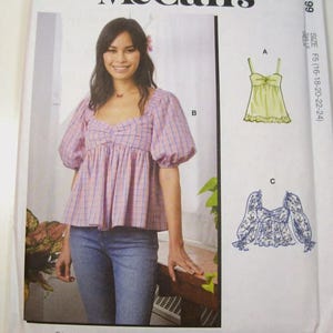 May include: A sewing pattern featuring a woman wearing a pink and blue plaid top with puffed sleeves and blue jeans. The pattern includes illustrations of a yellow top and a blue floral top. The pattern is for sizes 16-24.
