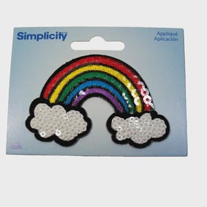Può includere: Una toppa applicata a forma di arcobaleno del marchio Simplicity, caratterizzata da un arco multicolore con strisce rosse, gialle, verdi, blu e viola, accentuato da paillettes argentate. La toppa include due nuvole bianche alla base, anch'esse decorate con paillettes.