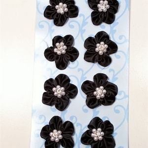 Könnte beinhalten: Eine Blatt mit acht schwarzen Stoffblumen mit weißen Perlenakzenten und einem Strassstein in der Mitte. Die Blumen befinden sich auf einem blauen Hintergrund mit einem weißen Wirbelmuster. Das Blatt zeigt unten links "8pc 6/oz" an.