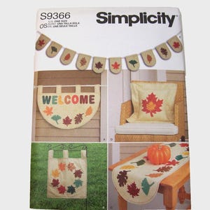 Puede incluir: Patrón de Simplicity con decoración otoñal para el hogar. Incluye una pancarta, un letrero de bienvenida, un cojín y un camino de mesa, todos adornados con diseños de hojas coloridas. La pancarta y el letrero de bienvenida tienen la palabra "WELCOME".