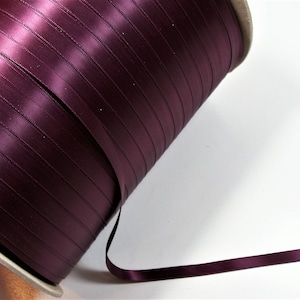 Puede incluir: Un carrete de cinta de satén burdeos oscuro. La cinta está bien enrollada en un núcleo de cartón, con un trozo de cinta desenrollada y arrastrando hacia la derecha. La cinta tiene una textura suave y brillante.