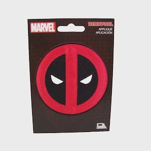 Puede incluir: Parche termoadhesivo de Deadpool sobre cartón. El parche presenta el diseño icónico de la máscara del personaje: un círculo rojo dividido por una línea roja vertical, con formas de ojos negros. El logotipo de Marvel es visible.