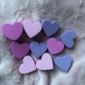 Puede incluir: Una colección de diez esponjas en forma de corazón en varios tonos de morado y rosa. Las esponjas están dispuestas en un patrón aleatorio sobre una superficie suave, peluda y de color morado claro.