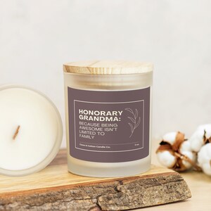 A candle with a mauve label that reads, "Honorary Grandma: Because being awesome isn't limited to family," rests on a wooden plank with its bamboo lid on. Another candle is on its side, showing creamy wax and a wood wick.