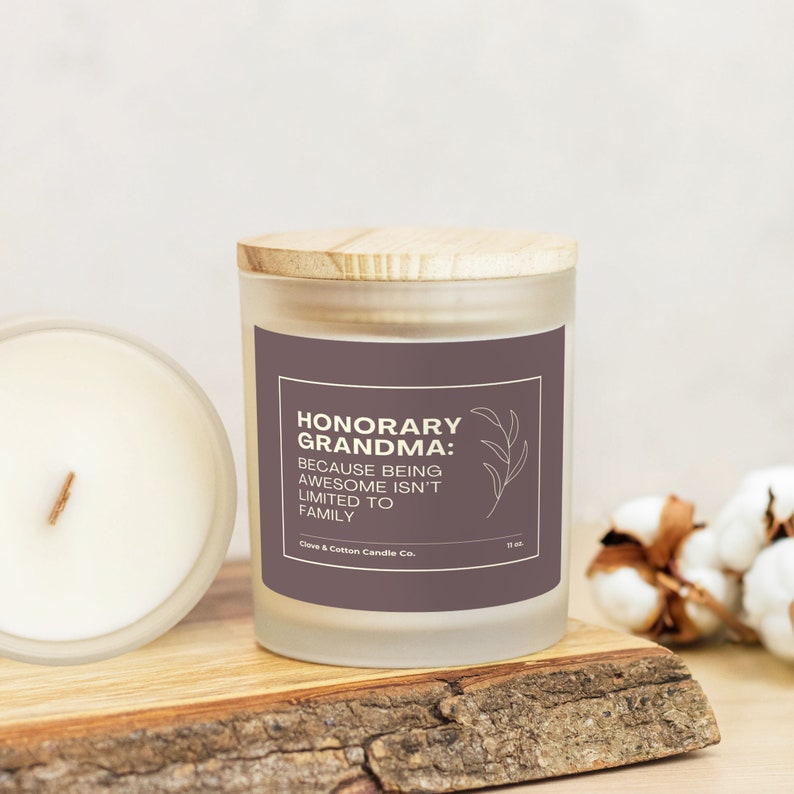 A candle with a mauve label that reads, "Honorary Grandma: Because being awesome isn't limited to family," rests on a wooden plank with its bamboo lid on. Another candle is on its side, showing creamy wax and a wood wick.