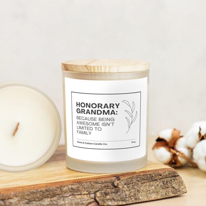 A candle with a white label that reads, "Honorary Grandma: Because being awesome isn't limited to family," rests on a wooden plank with its bamboo lid on. Another candle is on its side, showing creamy wax and a wood wick.