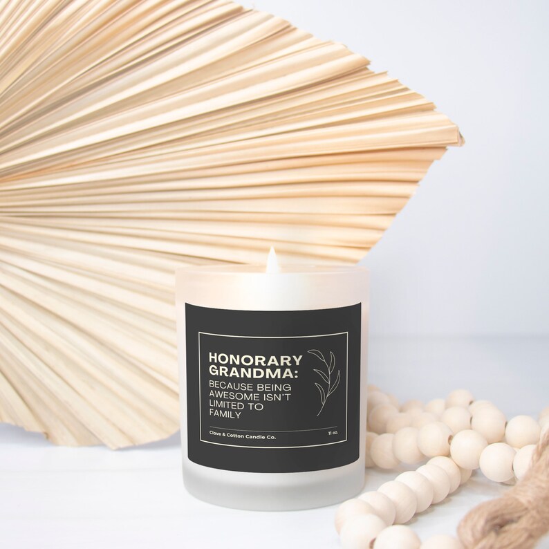 A candle with a grey label that reads, "Honorary Grandma: Because Being Awesome Isn't Limited to Family" burns in front of a cream-colored fan