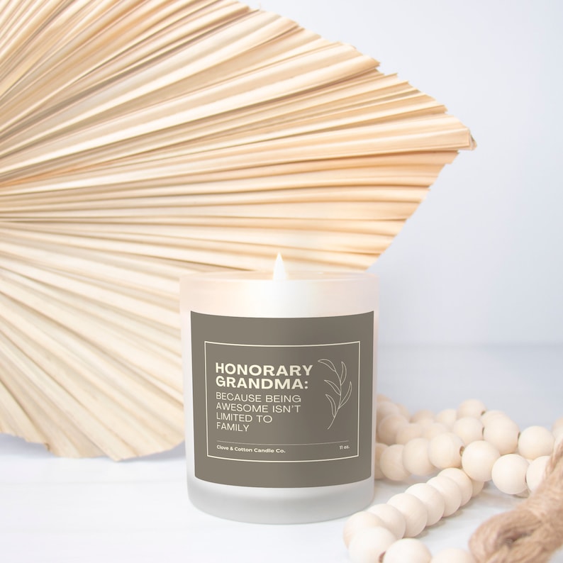 A candle with a brown label that reads, "Honorary Grandma: Because Being Awesome Isn't Limited to Family" burns in front of a cream-colored fan