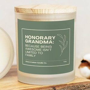 A candle with a sage label that reads, " Honorary Grandma: Because being awesome isn't limited to family," rests on a wooden plank with its bamboo lid on. Another candle is on its side, showing creamy wax and a wood wick.