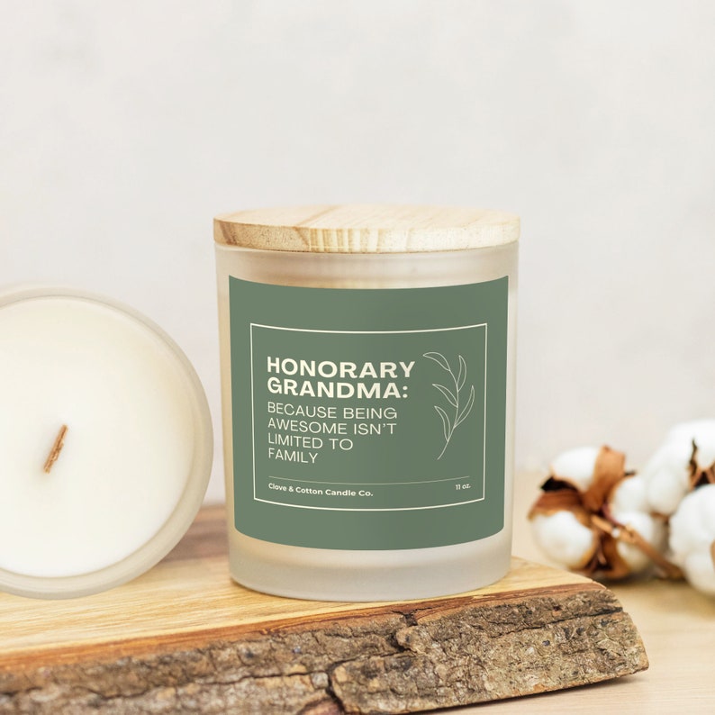 A candle with a sage label that reads, "Honorary Grandma: Because being awesome isn't limited to family," rests on a wooden plank with its bamboo lid on. Another candle is on its side, showing creamy wax and a wood wick.