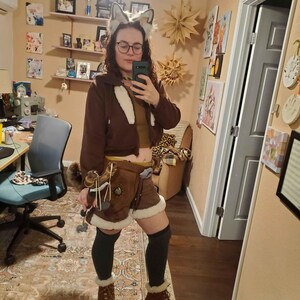May include: A brown hooded jacket with white trim, paired with brown shorts featuring faux fur trim. The outfit includes a pair of brown boots with faux fur lining, and a set of gray wolf ears. The outfit is completed with black thigh-high socks.