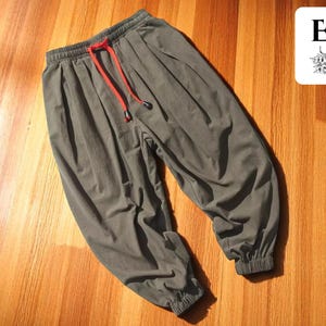 May include: Olive green pants with a red drawstring. The pants have an elastic waistband and cuffs. The pants are made of a soft material and are displayed on a wooden surface. A white square with the letters "EE" is in the upper right corner.