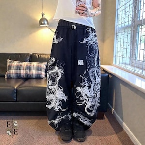 Könnte beinhalten: Schwarze Hose mit weitem Bein und weißem Drachendesign. Die Hose hat einen Kordelzug in der Taille und eine Cargotasche. Das Design ist aufwendig und bedeckt den größten Teil der Hose. Getragen mit schwarzen Schuhen.