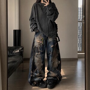 Könnte beinhalten: Ein dunkelgraues, zerfetztes Langarmshirt und eine weite, zerrissene Jeans. Die Jeans hat ein verwaschenes, abgenutztes Aussehen mit mehreren Rissen und Flicken. Das Outfit wird mit schwarzen Turnschuhen und einer silbernen Kette kombiniert.