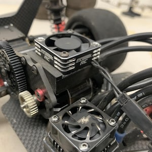 Op de afbeelding: Close-up van een zwarte elektromotor met twee koelventilatoren. De ventilatoren zijn met zilveren schroeven op de motor gemonteerd. De motor is verbonden met een zwarte draad. De tekst "ECRRC" is zichtbaar op de behuizing van de ventilator.