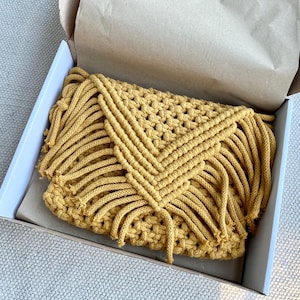 May include: A mustard yellow macrame clutch bag featuring a triangular flap with intricate knotted cord patterns and long fringe detailing along the edges, presented inside an open cardboard shipping box lined with brown tissue paper.