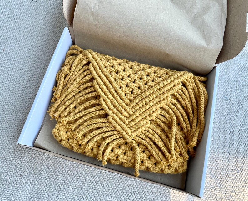 May include: A mustard yellow macrame clutch bag featuring a triangular flap with intricate knotted cord patterns and long fringe detailing along the edges, presented inside an open cardboard shipping box lined with brown tissue paper.