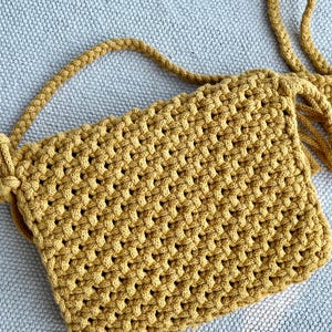 May include: A mustard yellow crossbody bag crafted with a dense macrame knot pattern. The bag features a rectangular body with a matching braided cord strap and decorative fringe details extending from the side attachment point of the strap.