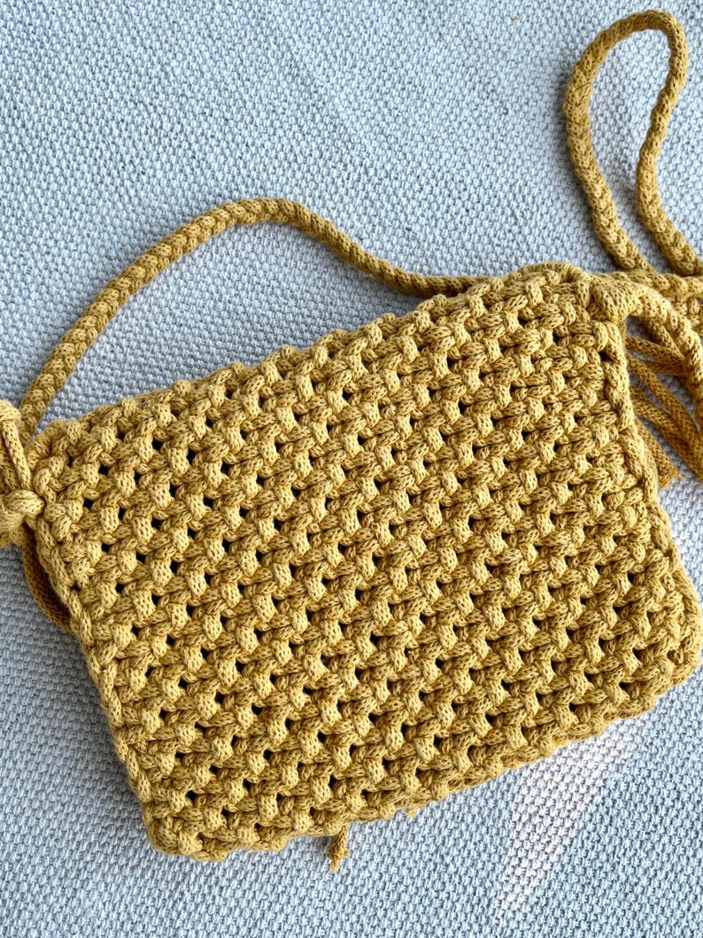 May include: A mustard yellow crossbody bag crafted with a dense macrame knot pattern. The bag features a rectangular body with a matching braided cord strap and decorative fringe details extending from the side attachment point of the strap.