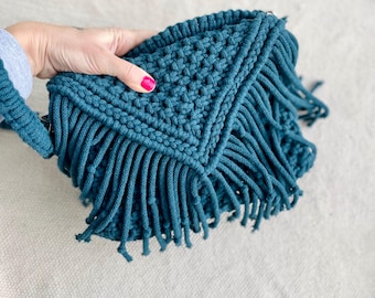 May include: A teal blue crocheted bag with a fringe detail. The bag is made with a macrame technique and has a triangular shape.