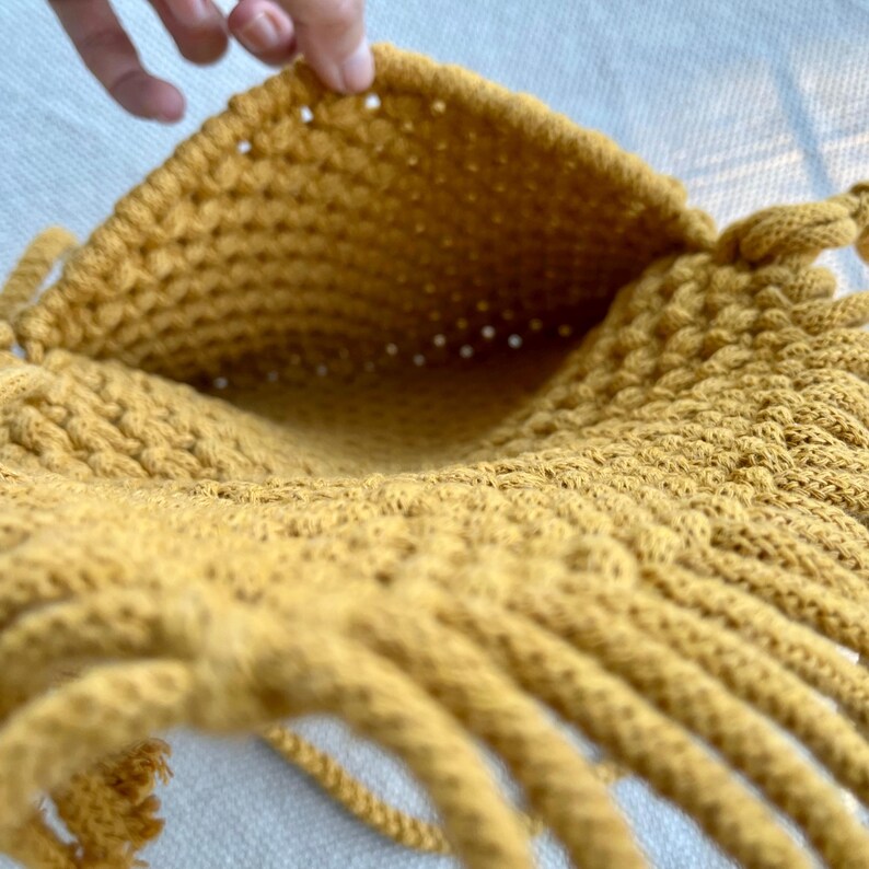 May include: A mustard yellow macrame bag featuring a dense, knotted cord construction with a wide, open top. The exterior displays a textured, woven pattern created by interconnected loops of thick cotton cord, forming a deep, hollow interior space.