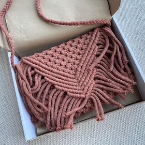 May include: A dusty rose macrame crossbody bag featuring a triangular flap with intricate knotted patterns and long fringe detailing. The bag includes a braided shoulder strap and is constructed from thick cotton cord in a textured design.