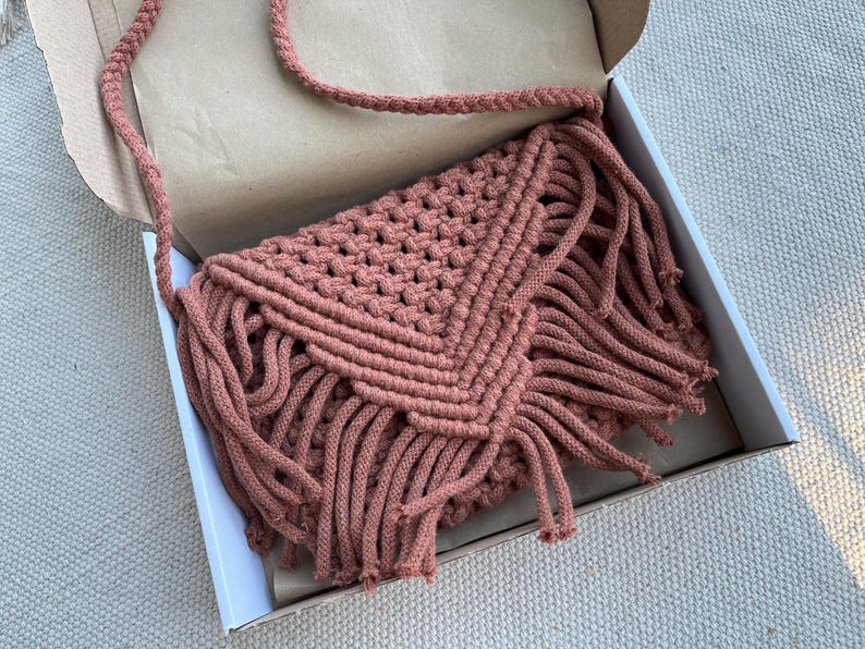 May include: A dusty rose macrame crossbody bag featuring a triangular flap with intricate knotted patterns and long fringe detailing. The bag includes a braided shoulder strap and is constructed from thick cotton cord in a textured design.