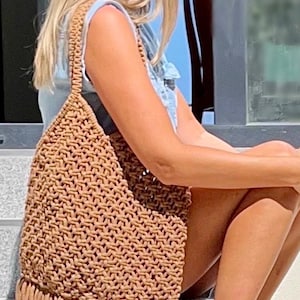 May include: A tan macrame shoulder bag featuring a triangular silhouette with a textured knotted pattern and a thick fringe trim along the bottom edge. The bag includes a single integrated shoulder strap made from the same knotted cord material.