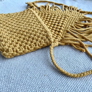 May include: A mustard yellow macrame crossbody bag featuring a dense, textured knot pattern on the main body and a decorative fringe trim along the bottom edge. The bag includes a long, braided cord strap attached to the top corners.