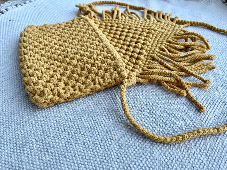 May include: A mustard yellow macrame crossbody bag featuring a dense, textured knot pattern on the main body and a decorative fringe trim along the bottom edge. The bag includes a long, braided cord strap attached to the top corners.