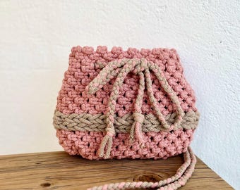 May include: A pink and beige crocheted bag with a drawstring closure and a long strap. The bag is made of a soft, textured yarn and has a bohemian style.