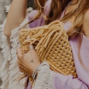 May include: A mustard yellow macrame clutch bag featuring a textured knotted cord pattern with fringe details along the top edge. The bag is constructed from thick cotton rope and displays a structured rectangular shape with a woven grid design.