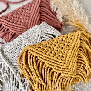 May include: Three envelope-style macrame bags in terracotta, light gray, and mustard yellow. Each bag features a triangular flap with a woven knot pattern and long, dangling fringe along the bottom edge, showcasing a textured cord construction.