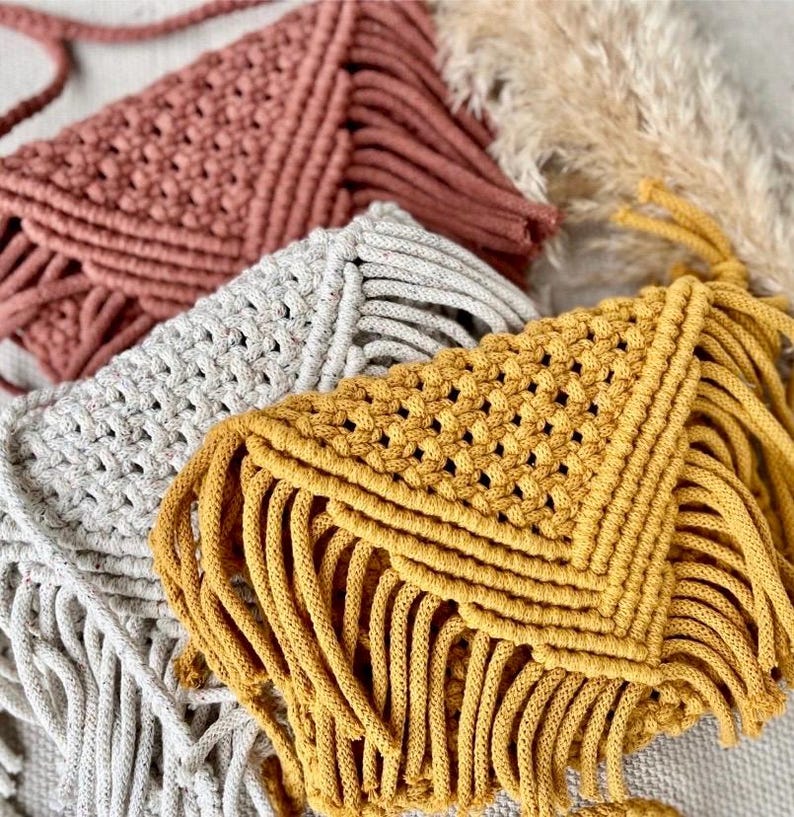May include: Three envelope-style macrame bags in terracotta, light gray, and mustard yellow. Each bag features a triangular flap with a woven knot pattern and long, dangling fringe along the bottom edge, showcasing a textured cord construction.