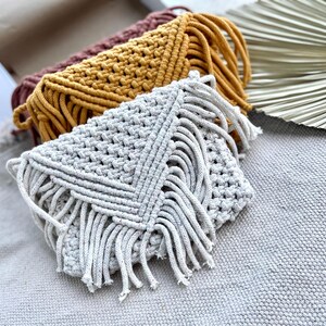 May include: Three macrame clutch bags in light gray, mustard yellow, and dark mauve. Each envelope-style bag features a textured knotted cord pattern with a triangular flap and long fringe detailing along the edges, showcasing intricate fiber craft.
