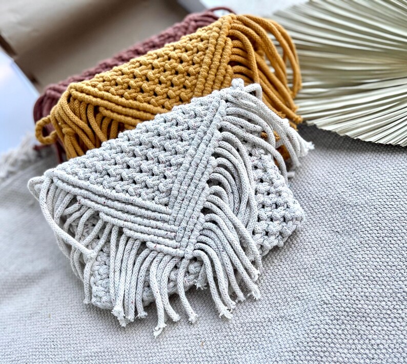 May include: Three macrame clutch bags in light gray, mustard yellow, and dark mauve. Each envelope-style bag features a textured knotted cord pattern with a triangular flap and long fringe detailing along the edges, showcasing intricate fiber craft.