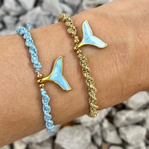 May include: Two braided bracelets with gold accents and blue enamel whale tail charms. The bracelets are on a person's wrist.