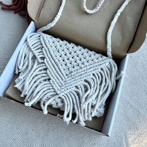 May include: An off-white macrame crossbody bag featuring a triangular flap, intricate knotted cord patterns, and long fringe detailing along the bottom edges. The bag includes a braided shoulder strap and is presented inside an open cardboard box.